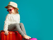 © Ilshat - Little cute girl in straw hat and sunglasses sitting on suitcases, portrait of beautiful child going on a journey, studio shot over isolated blue isolated.
