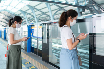 two asian women using smart phone and standing at the gate of platform of sky train station. They keep social distancing to avoid covid-19 or coronavirus infection. new normal lifestyle concept