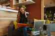 © Dragica - A beautiful caucasian business woman is sitting in a cafe, talking on the phone and working on a laptop. Work in a cafe