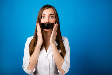 Girl doctor in a white coat with black tape over her mouth on a blue background close-up, silence and non-disclosure of medical secrets