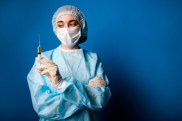 Doctor in blue uniform standing on a blue background in a medical protective mask holding a vaccination syringe in his hand