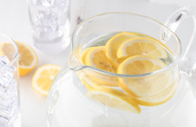 A Close Up View Of A Jug Of Lemon Water With Slices Of Lemon And Two Glasses Of Ice Ready For Pouring.