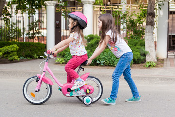 children learning to drive a bicycle on a driveway outside. a little girl teaches her sister to ride