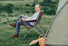 Young Man Freelancer Sitting On Chair And Relaxing In Front Of Tent At Camping Site In Forest Or Meadow. Outdoor Activity In Summer. Adventure Traveling In National Park. Leisure, Vacation, Relaxation