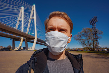 A young man stands against a cable-stayed engineering bridge in a medical mask