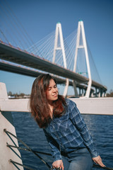 A brunette girl with long hair in a blue shirt stands against the background of a cable stayed bridge on the embankment