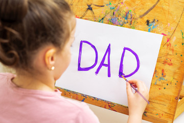 Poster - Little girl is drawing handwritten purple word Dad greeting card by watercolors on white paper on easel. Father's Day and family concept.