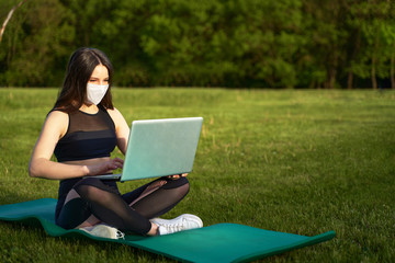 A girl in a medical mask sits with a laptop on the lawn