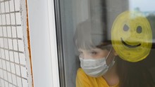Child, Little Girl In A Medical Mask Sad Looking Out The Window. Face Emoticon Symbol Is Painted On The Window. Isolated People, Childs Quarantined At Home, Virus Epidemic. Shows Pozitiv Emotions.