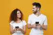 © ViDi Studio - Smiling young couple friends european guy african american girl in white t-shirts isolated on yellow background. People lifestyle concept. Using mobile phone typing sms message, looking at each other.