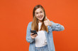© ViDi Studio - Funny young woman girl in casual denim clothes posing isolated on bright orange wall background studio portrait. People lifestyle concept. Mock up copy space. Using mobile phone, showing thumb up.