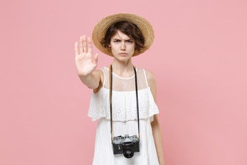 Displeased young tourist woman in summer white dress hat with photo camera isolated on pink background. Traveling to travel weekend getaway. Air flight journey concept. Showing stop gesture with palm.