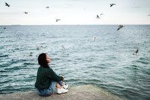  A Young Girl Sits On A Pier By The Sea In Windy Weather And Reflects On Life. Seagulls Fly Over The Water. Loneliness. Calmness And Peace. Charge Of Inspiration. Sea Breeze