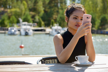 Young beautiful Asian tourist woman using phone at restaurant by the pier