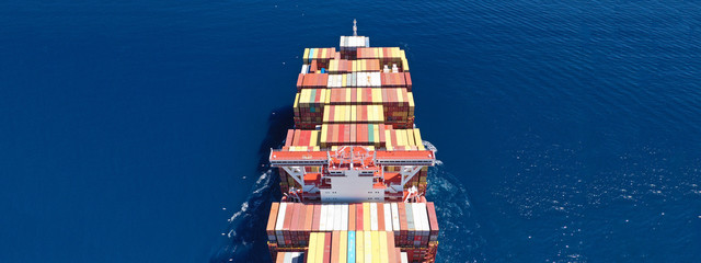 Aerial ultra wide photo of Container cargo Ship carrying load in truck-size colourful containers in deep blue open ocean sea 