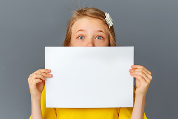 A teenage girl in yellow clothes on a grey background holds a blank white poster for the concept insert