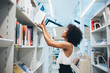 Young african american woman taking book from high shelf in library searching literature for self education in college, dark skinned curly female librarian putting literature in order during work