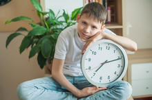 Teenager Boy In A White T-shirt Sits On A Table In A Children's Room With Legs Crossed, Leaned On A Large Silver Watch. Dreamy, Pensive, Smiling.