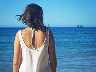 Mujer de espaldas mira al horizonte sobre el mar donde se ven dos pequeños islotes, luz de mañana