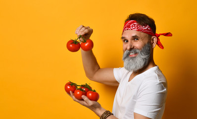 Wall Mural - Middle-aged guy in red bandana, white t-shirt and bracelet. He is smiling, showing red tomatoes, posing on orange background