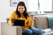 © Syda Productions - internet bank, online shopping and technology concept - happy smiling asian young woman in yellow sweater sitting on sofa with tablet pc computer and credit card at home