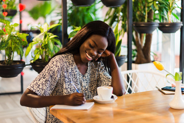 Portrait of beautiful young african woman sitting at cafe and writing notes