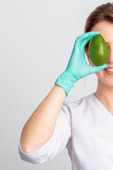 Wall Mural - Half face portrait of woman cosmetologist or doctor covering her eye by an avocado on white background. Healthy and beauty concept.