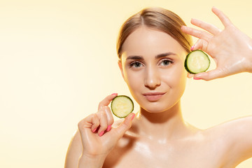 Wall Mural - Close up of beautiful young woman with cucumber slices on yellow background. Concept of cosmetics, makeup, natural and eco treatment, skin care. Shiny and healthy skin, fashion, healthcare.