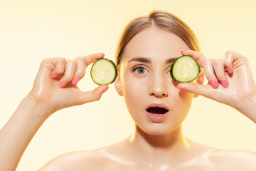 Wall Mural - Close up of beautiful young woman with cucumber slices on yellow background. Concept of cosmetics, makeup, natural and eco treatment, skin care. Shiny and healthy skin, fashion, healthcare.