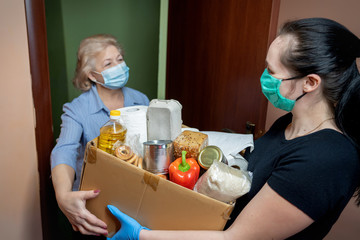Canvas Print - Volunteer young female in medical mask and gloves handing an senior woman a box with food. Donation, support people in quarantine, coronavirus