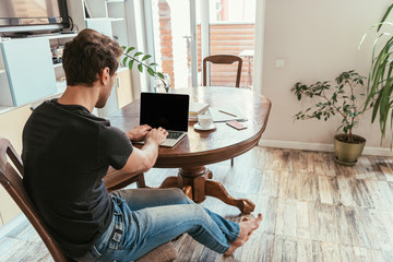 back view of young man typing on laptop with blank screen at home