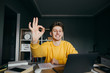 © bodnarphoto - Positive young man in an orange sweatshirt sits at a table at home with a computer and books, studies remotely, looks at the camera with a smile on face and shows the piles gesture OK.Studying at home