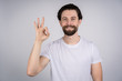 © Ivan - A young man with a beard in a white t-shirt standing on gray background, hand showing thumbs up OK