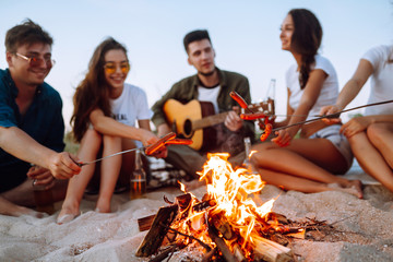 Fried sausages on bonfire. Group of young friends sitting on beach and fry sausages. Summer holidays, vacation, relax and lifestyle consept. Camping time.