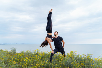 Two sportsmen practicing acro yoga together outdoors in the evening. Balancing girl legs up. Perfect flexibility. Athlete standing upside down. Holding a girl with one hand. Long shot