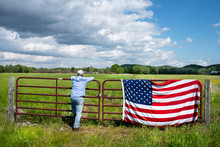 Farmer Leaning On Metal Fence In Grassy Agricultural Field, American Flag Draped Over Fence, Blue Sky White Clouds