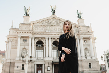 A beautiful young woman in a black dress with blond hair walks around the city with a bag with red flowers, filtered image, flare sunshine