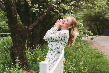 Portrait young carefree hippie girl in white floral dress have fun and smile on green countryside garden. Beautiful stylish blonde woman posing with wooden fence in summer park. Enjoy nature