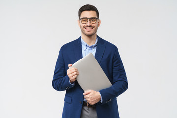 Smiling business man looking at camera through trendy glasses, holding closed laptop next to chest as if going to office, isolated on gray background