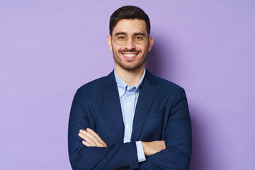 Portrait of handsome businessman in round eyeglasses standing in front of camera, looking with smile, holding arms crossed, ready to consult and support, isolated on purple background