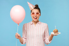 Happy Girl In Colored Glasses Celebrating Birthday, Wearing Special Hat, Holding Cake On Plate And Pink Balloon, Isolated On Blue Background