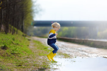 Mischievous preschooler child wearing yellow rain boots jumping in large wet mud puddle after rain. Kid playing and having fun in sunny spring day. Outdoors games for children.