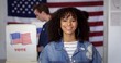 © vesperstock - MCU Young Hispanic woman in denim jacket with a new 'I Voted' sticker, smiling and standing in front of voters in polling booths with US flag in background
