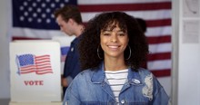 MCU Young Hispanic Woman In Denim Jacket With A New "I Voted" Sticker, Smiling And Standing In Front Of Voters In Polling Booths With US Flag In Background