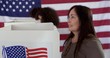© vesperstock - CU mature Caucasian woman thinking while casting votes at booth in polling station. Other voters and large US flag behind.