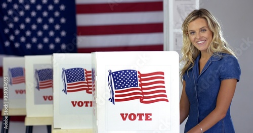 MS Young blonde woman casting vote at booths, looks up and smiles in polling station with US flag.