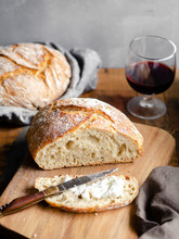 A Homemade Loaf Of Bread, Sliced And Buttered On A Wood Cutting Board Sitting Atop A Rustic Table With A Glass Of Red Wine In The Background