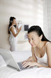 © ImageHit - Woman lying forward on bed using laptop, friend listening to music in the background