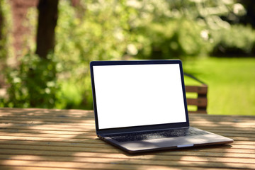 A laptop with an empty screen stands on a wooden table in the courtyard of a beautiful home garden, in Sunny weather.