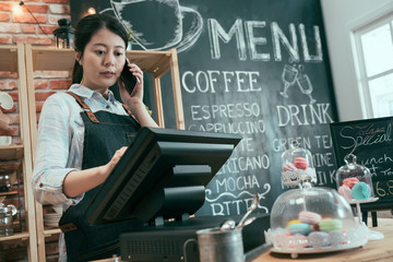 Wall Mural - young asian japanese woman staff working in cafe store. lady waitress talking on cellphone while customer taking order. female barista using point of sale terminal and mobile phone in coffee shop.